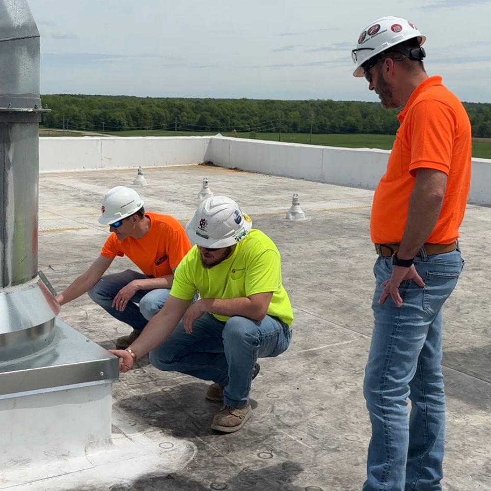 Quality_Body_CTta Mixer Expansion Two construction workers inspect a large pipe on a roof while a third worker looks on