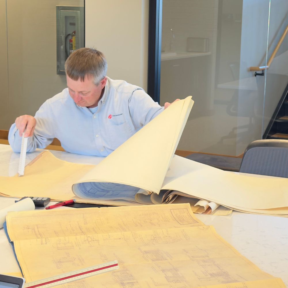 A man in a collared shirt examines a building plan while holding a ruler