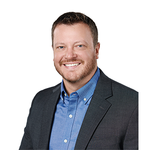 A smiling man with an auburn mustache and beard, a blue collared shirt, and a gray sports coat