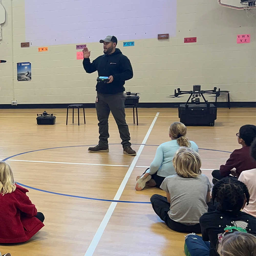 PKWY River Bend_901 NG_2024-12_Drone Pres_IMG_4405 A man with three drones behind him addresses a crowd of children