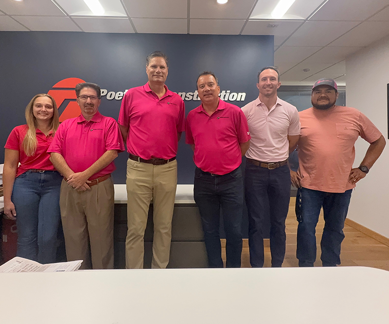 Five men and one woman wearing pink shirts in front of a blue wall with a Poettker Construction logo