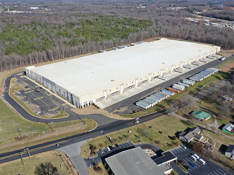 Aerial view of a large distribution center/warehouse with a white roof with a forest behind it and an industrial park in front