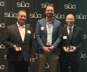 Three men holding two awards pose in front of a SLC3 step-and-repeat