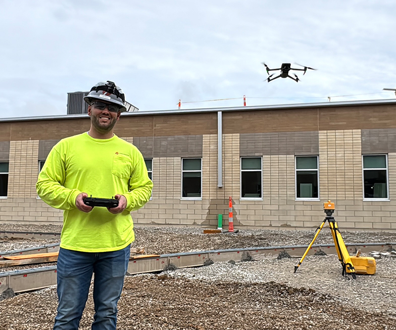 A man in a high-visibility shirt and hard hat holds a remote as a drone hovers a few feet away